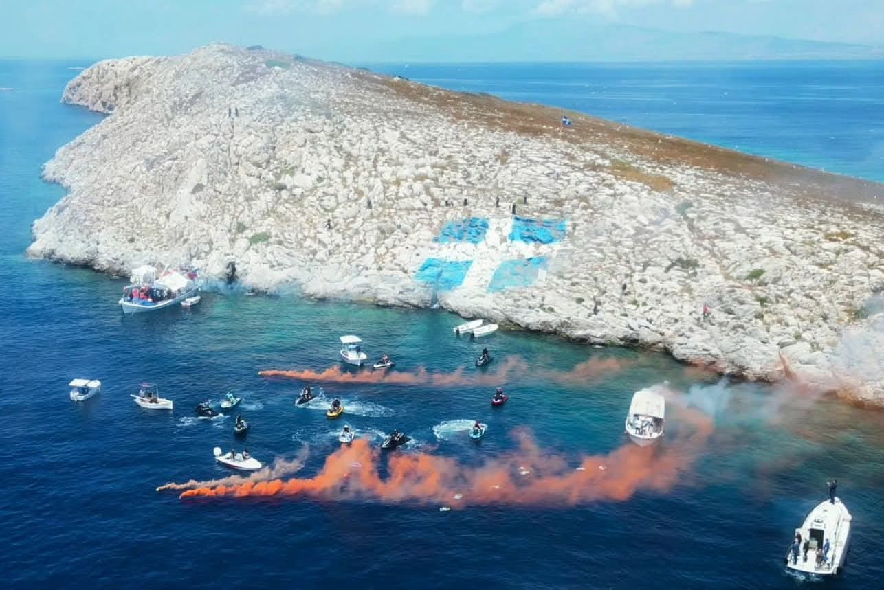 Από ψηλά: jet ski και σκάφη γύρω από βραχονησίδα με ζωγραφισμένη ελληνική σημαία, με πορτοκαλί καπνογόνα