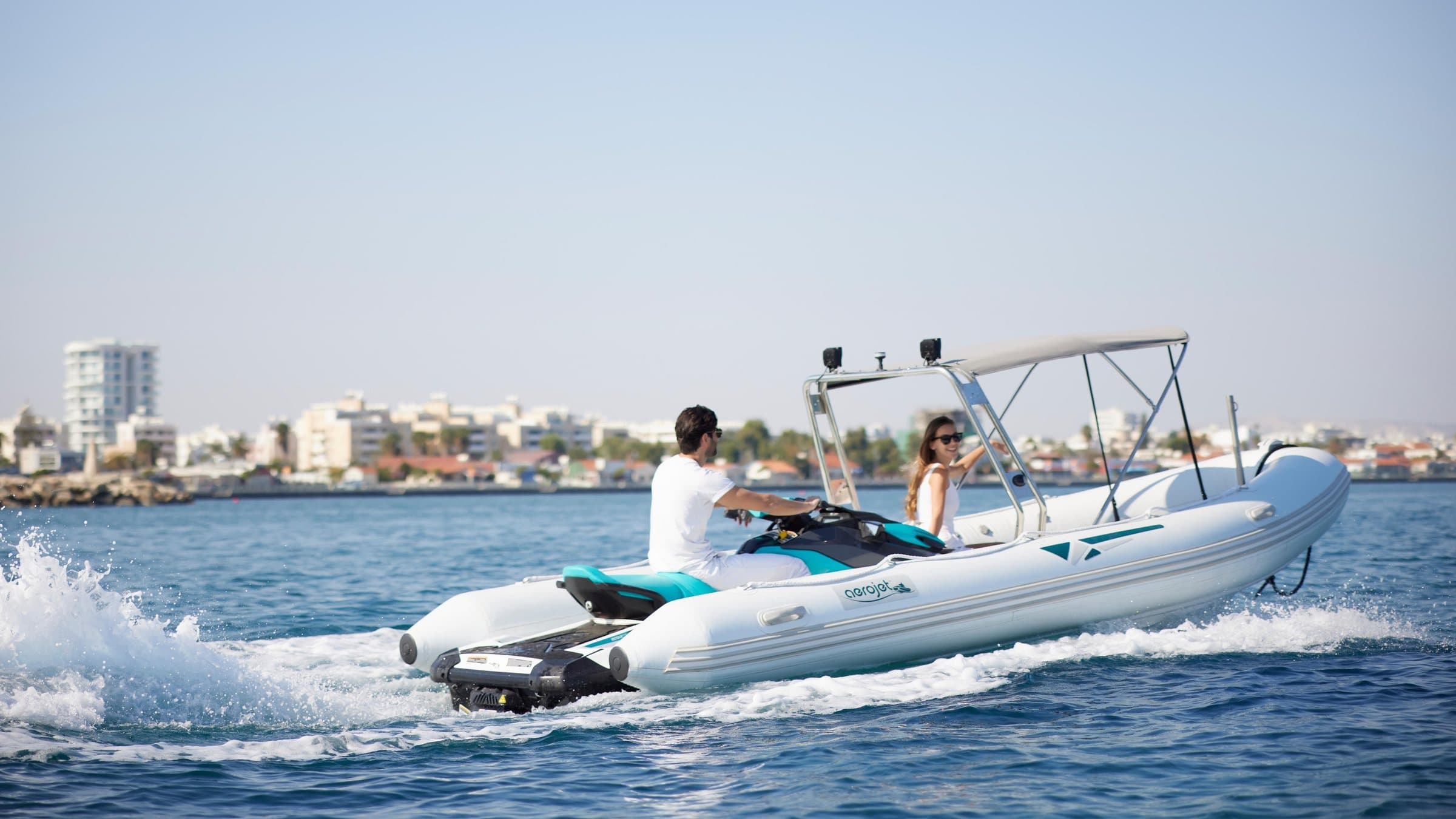 Το Aerojet 5.6 σε πλεύση με δύο επιβάτες και το jet ski κουμπωμένο στην πρύμνη, με την ακτή στο βάθος